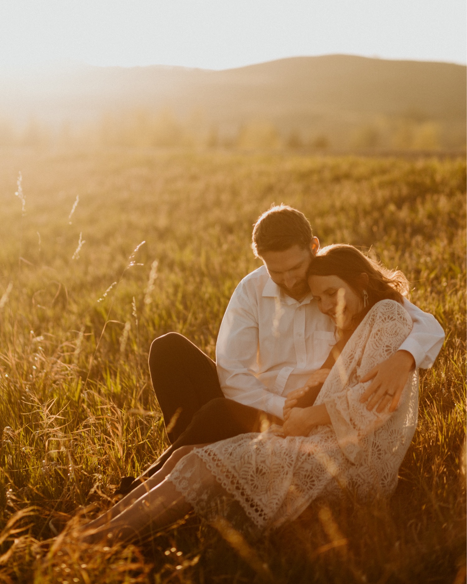 Maternity Family Photos in Calgary with mother and father dressed up for their pregnancy photoshoot with white flowy dress and baby boy in mothers belly near the mountains in Calgary Alberta during a beautiful sunset, maternity lifestyle photos in Calgary, casual lifestyle maternity photoshoot, baby photos