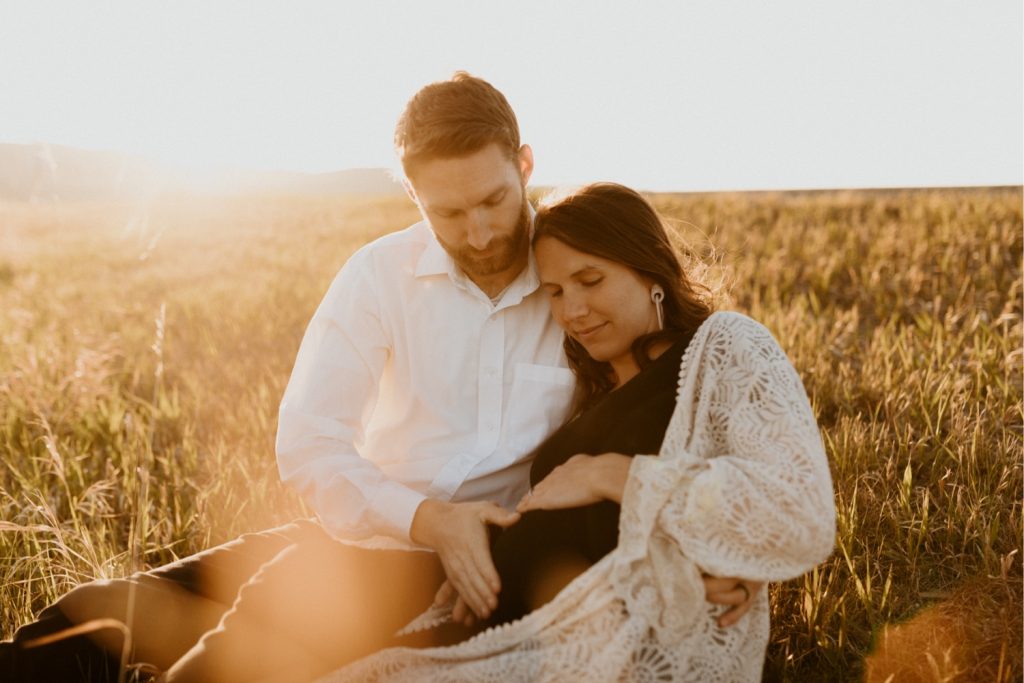 Maternity Family Photos in Calgary with mother and father dressed up for their pregnancy photoshoot with white flowy dress and baby boy in mothers belly near the mountains in Calgary Alberta during a beautiful sunset, maternity lifestyle photos in Calgary, casual lifestyle maternity photoshoot, baby photos